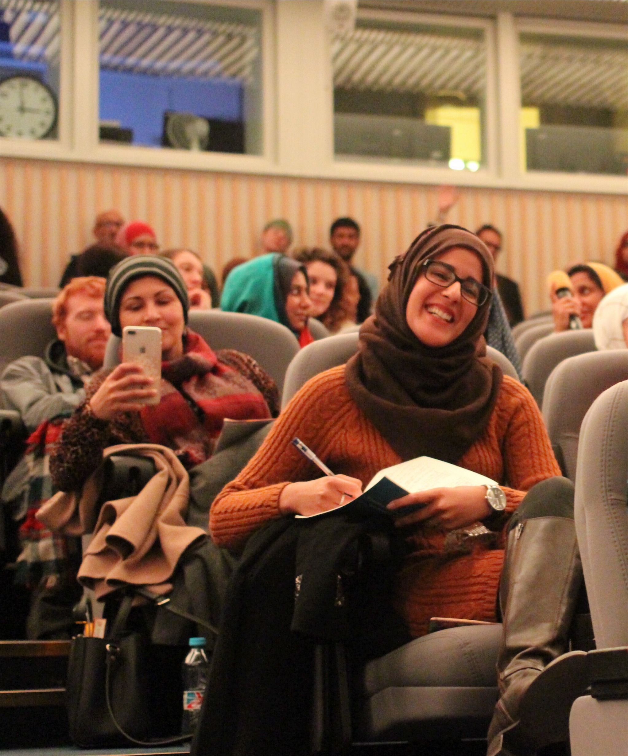 woman in a brown hijab, orange jumper and glasses laughing with a pen and notebook and sitting in a lecture theatre surrounded by other people.