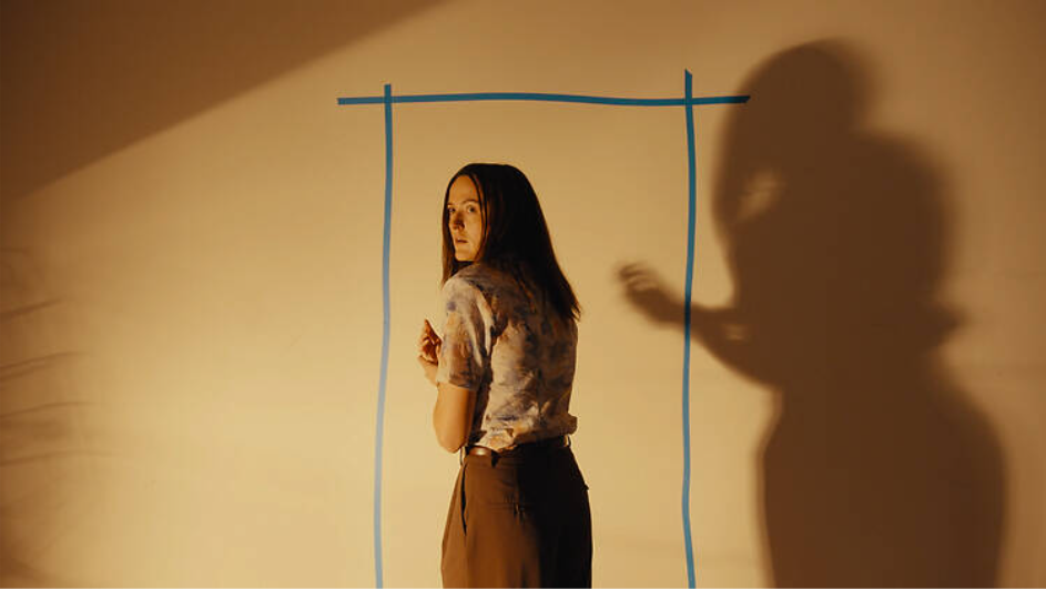 A still from the horror film Backrooms, showing a white woman looking back scared, with a wall in front of her with her shadow and a blue rectangle taped on it