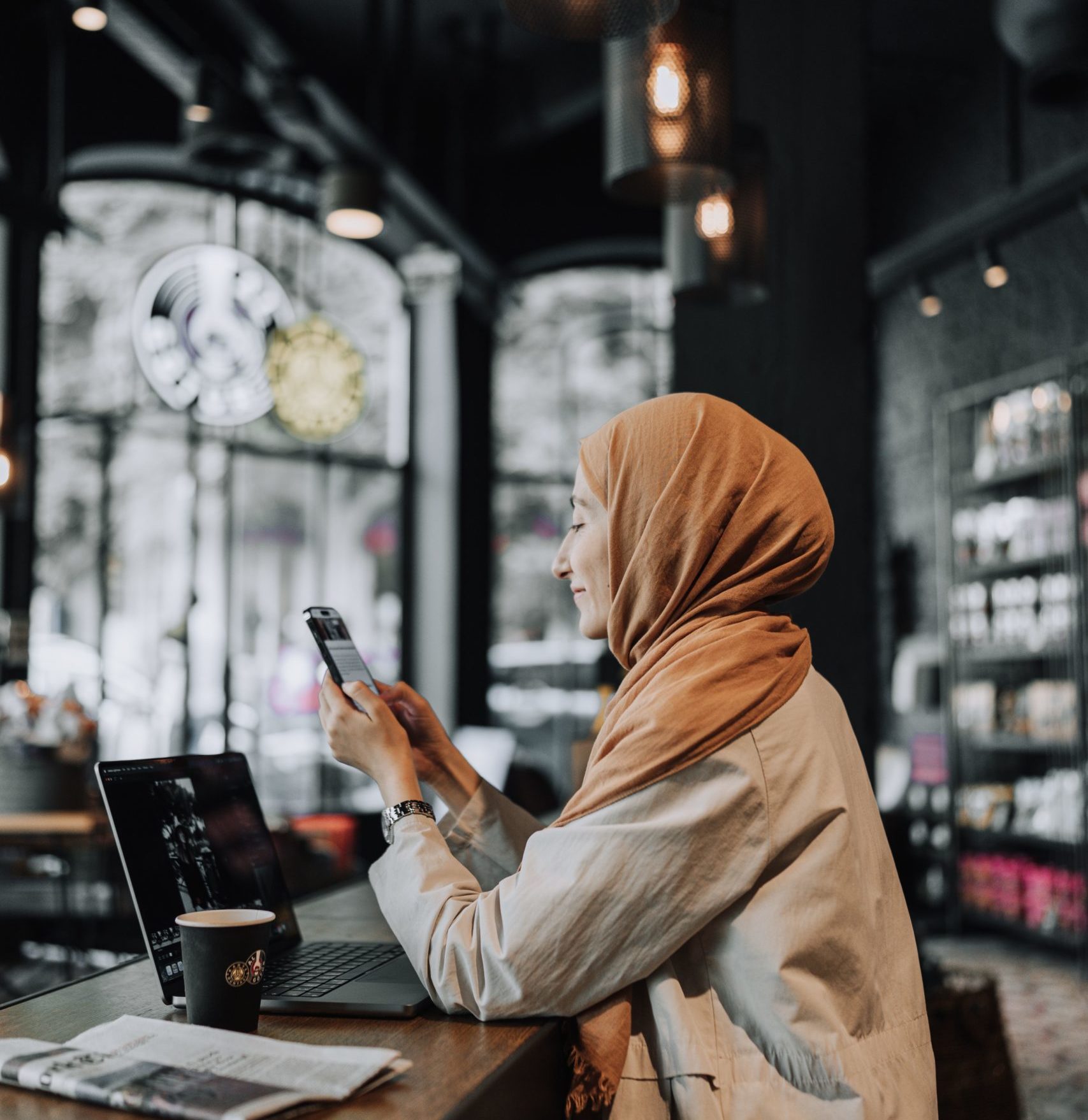 A hijabi woman sitting in cafe looking at her phone with a laptop open, cup of coffee and newspaper next to it. 
the evil eye and social media