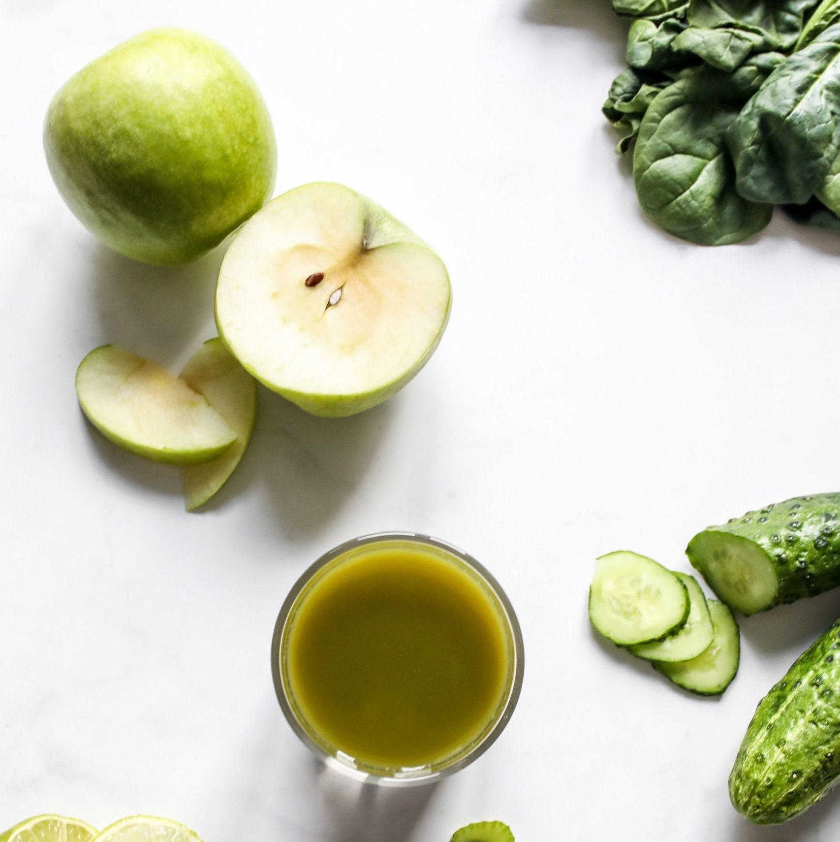 image shows a flatlay showing green juice in a glass with a full and half cut green apple, cucumber and spinach/kale around it